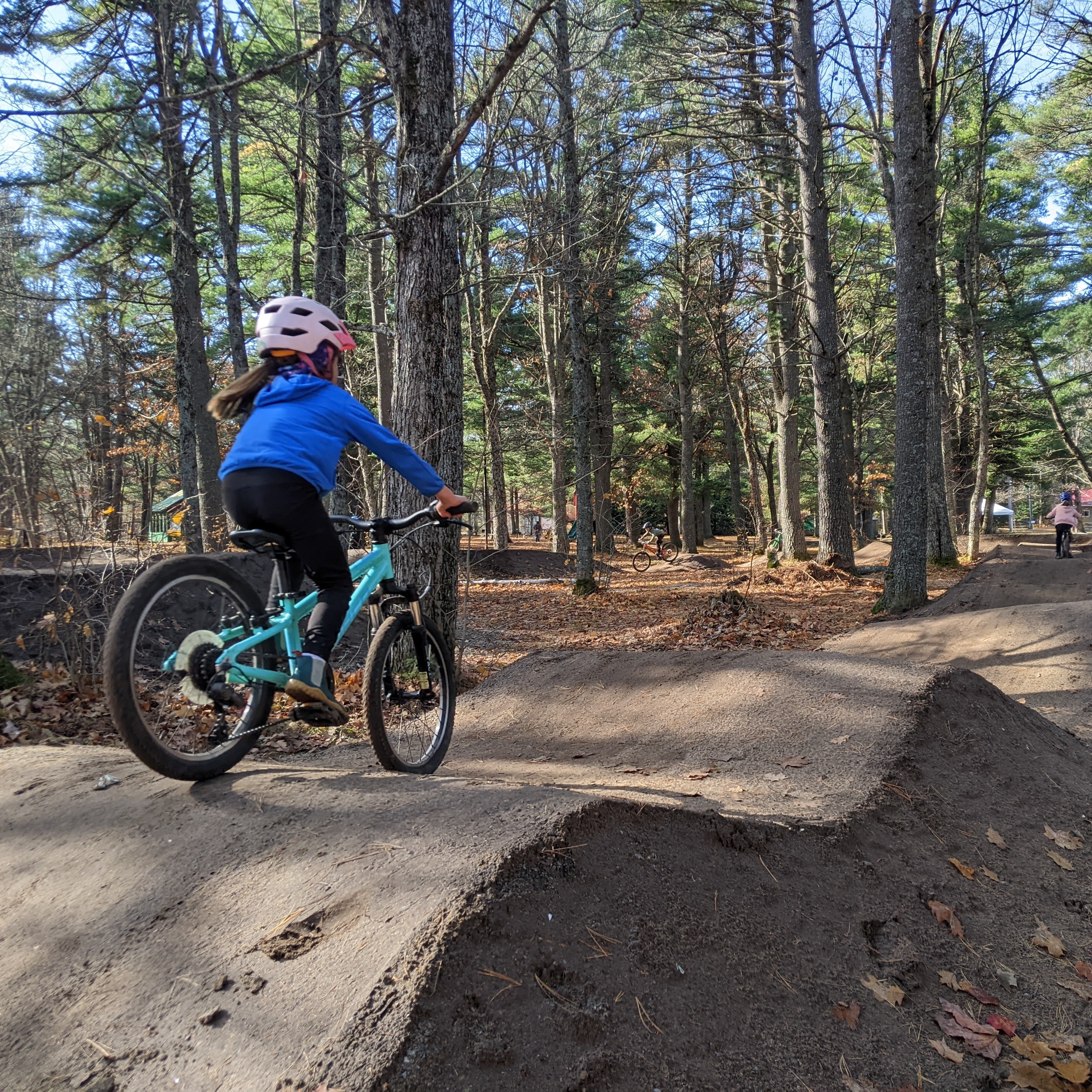 young girl rides a bicycle over a series of dirt ramps along a forest path