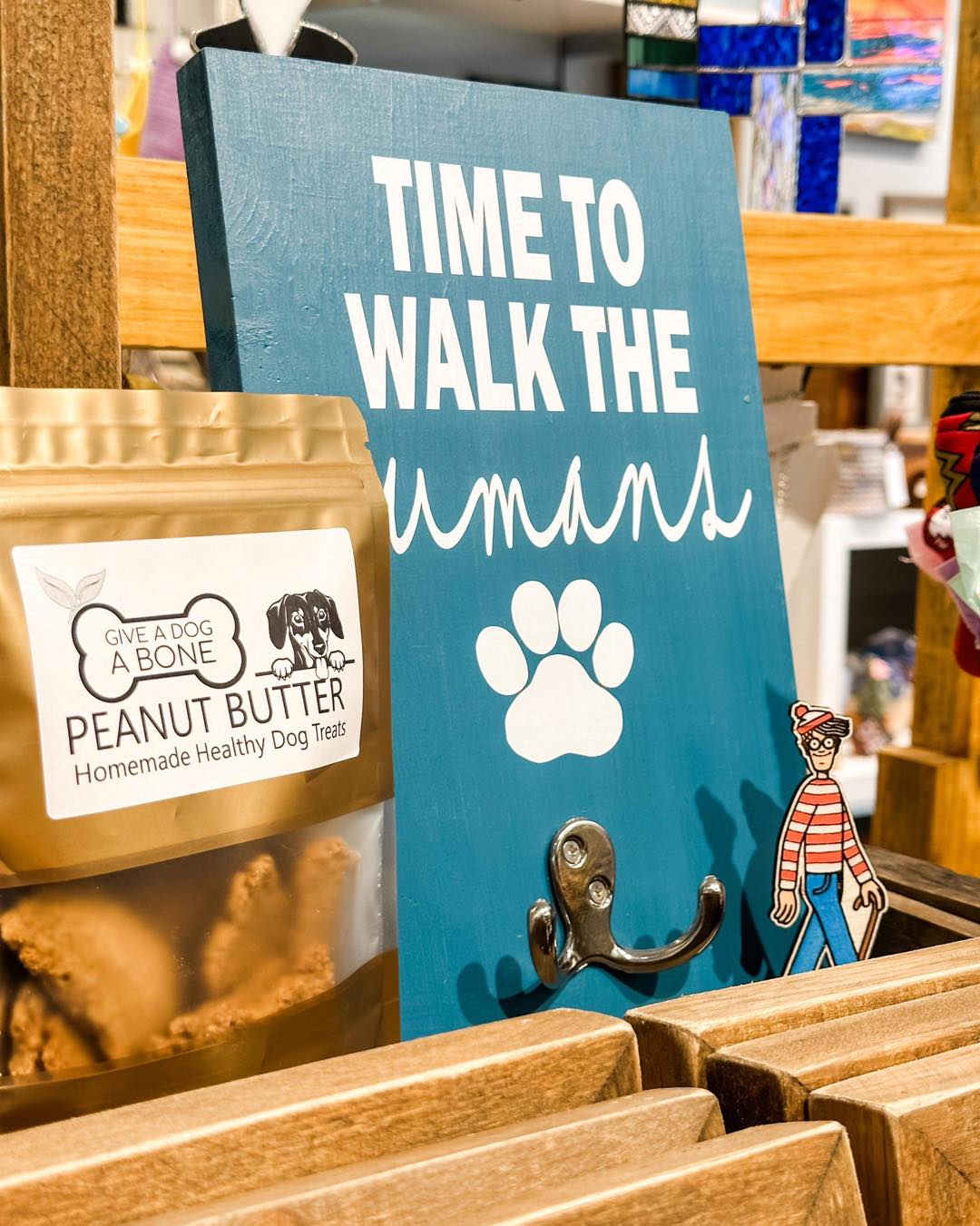a wooden display shelf showing a bag of home-made peanut butter dog treats, a turquoise wall plaque with a leash hook on it that says "time to walk the humans" in white, and a small Where's Waldo toy hidden next to that.