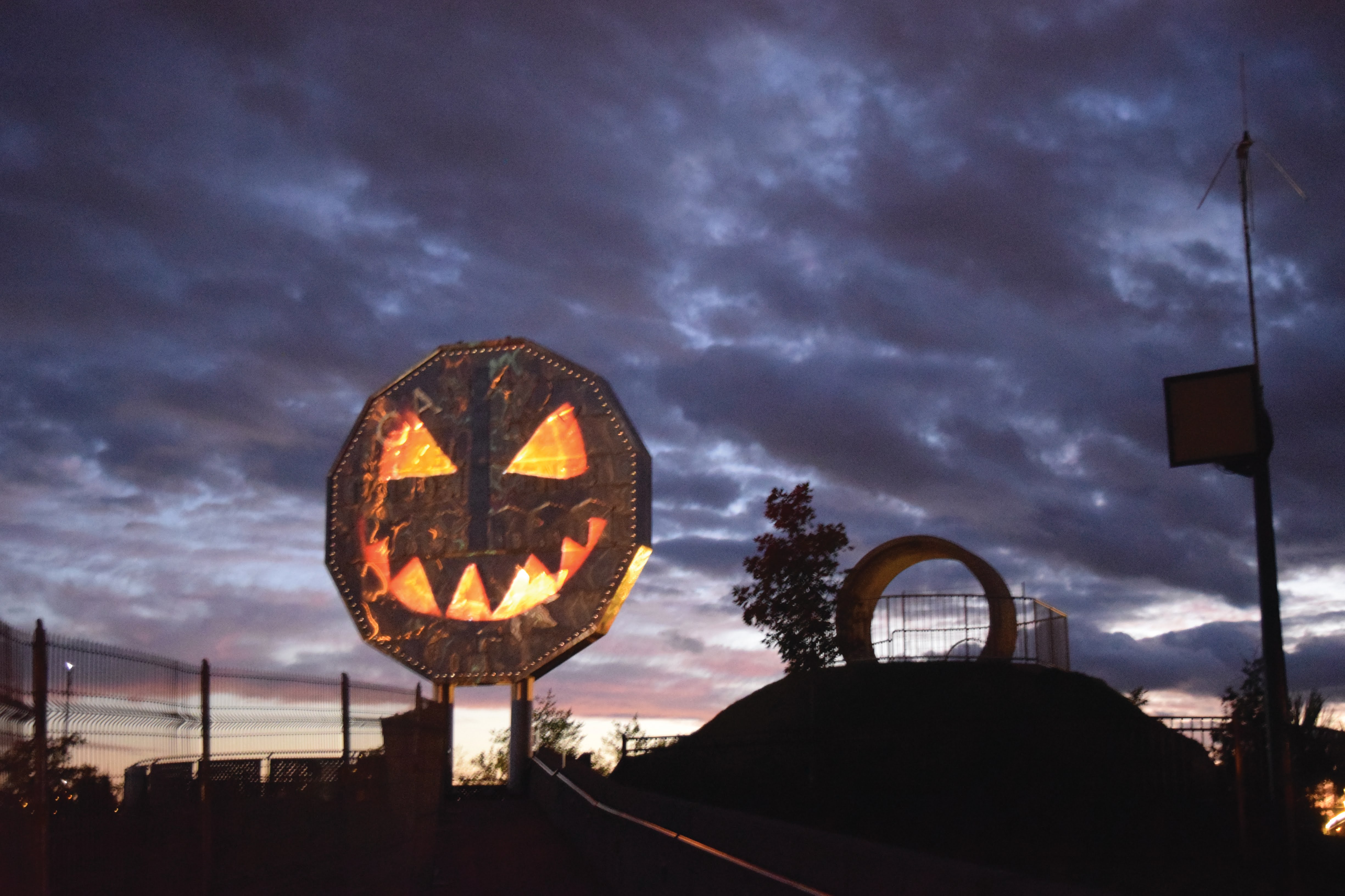 The Sudbury Big Nickel Monument in front of a dusky, clouded sky, with the face of a Jack-o-lantern projected onto the nickel in orange light.