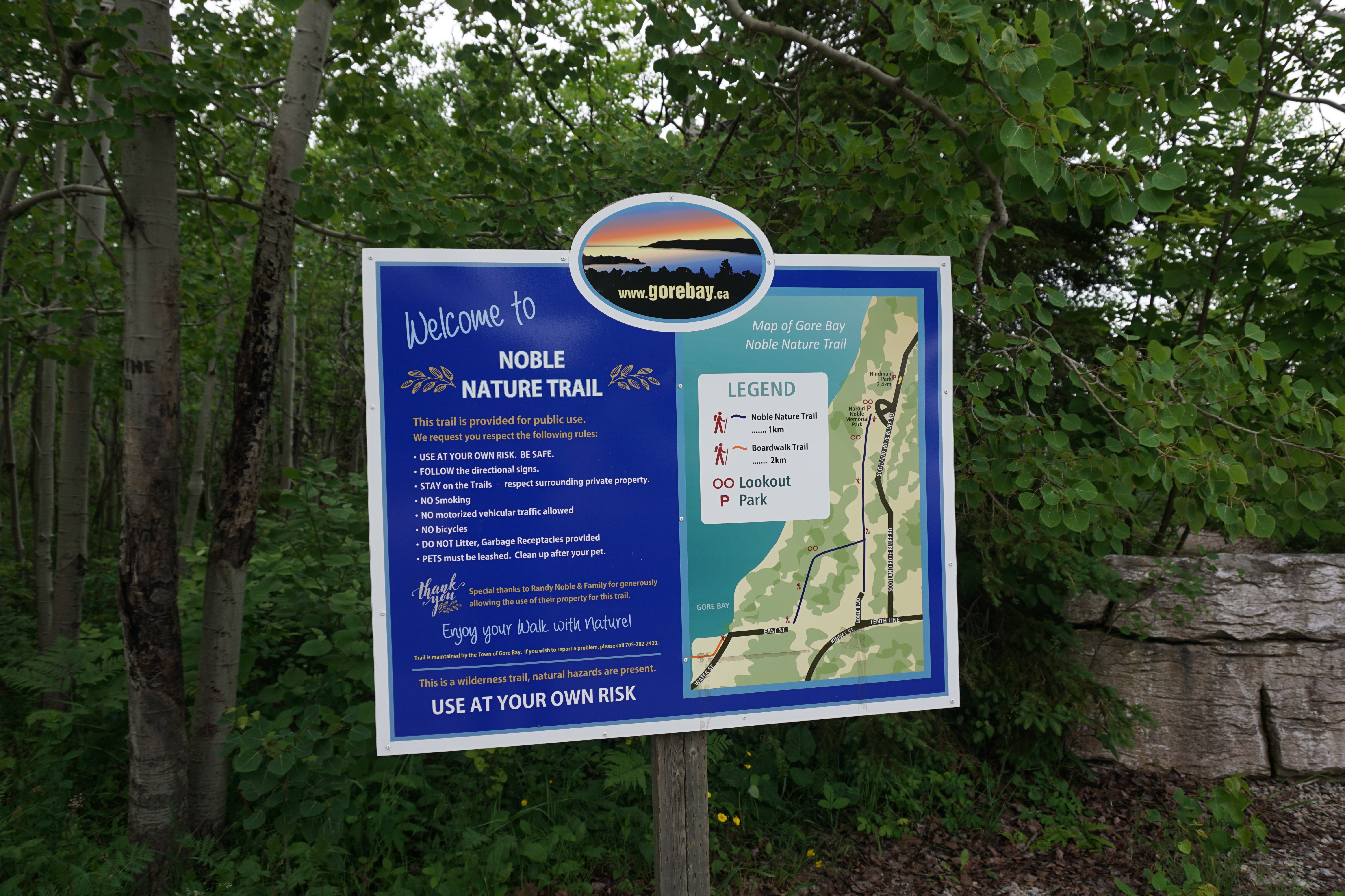 a blue park informational sign for" The Noble Nature Trail" near Gore Bay, with lush green trees in the background.