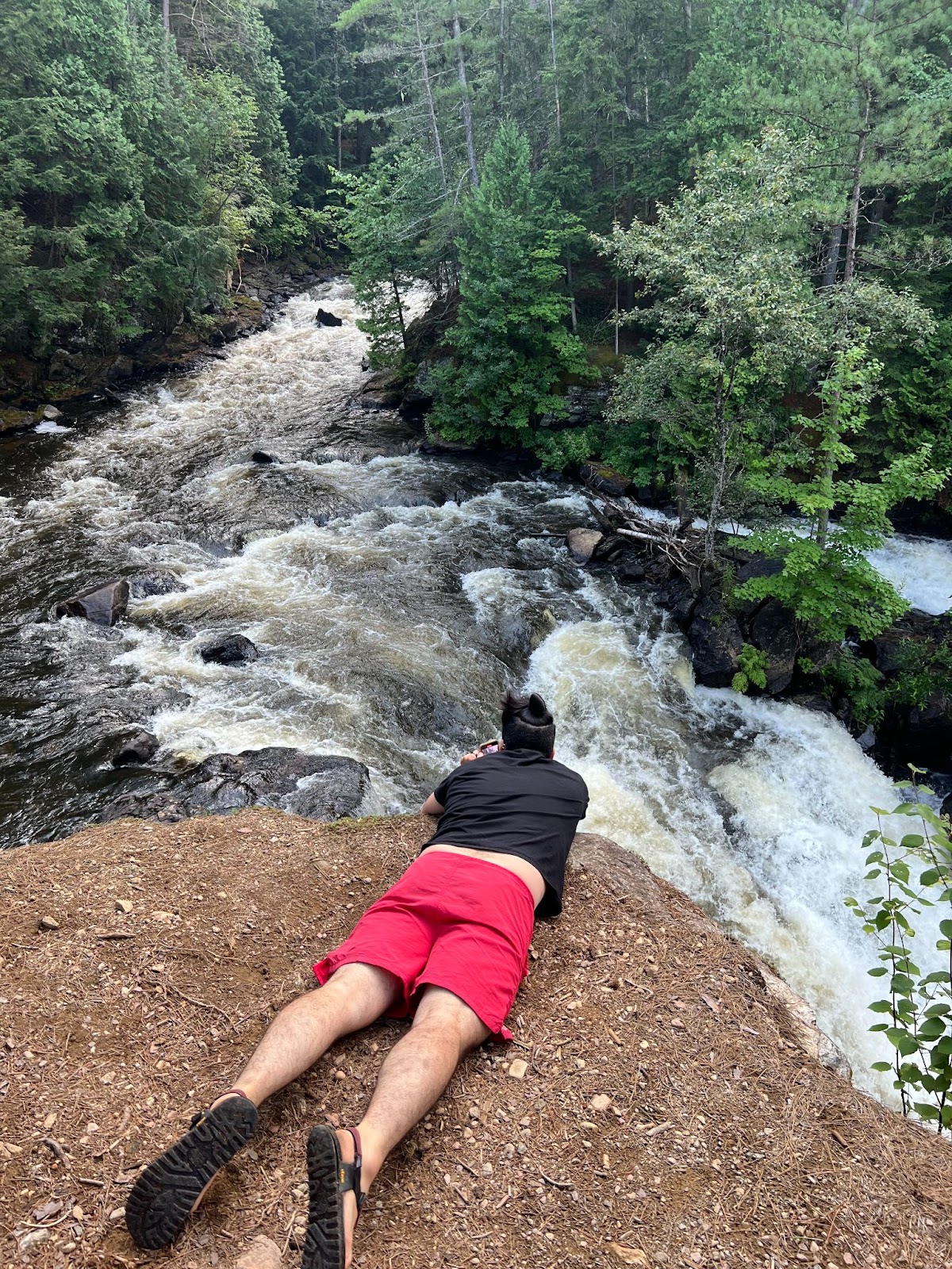 Eau Claire Gorge; a man lays on his belly looking down into a high, tree-filled gorge with a raging river and waterfall through it.