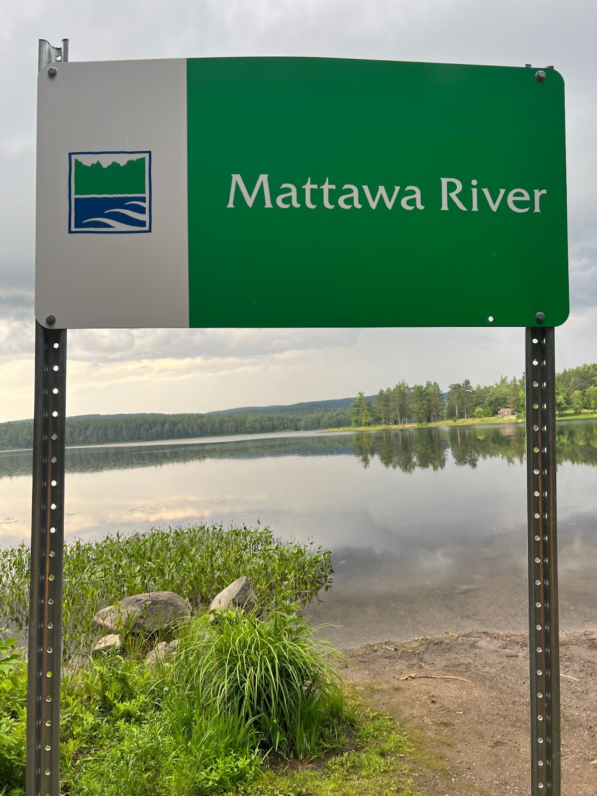 a green and white Ontario Parks sign that reads "Mattawa River", in front of a wide, glassy river with thick green forest on the distant bank.