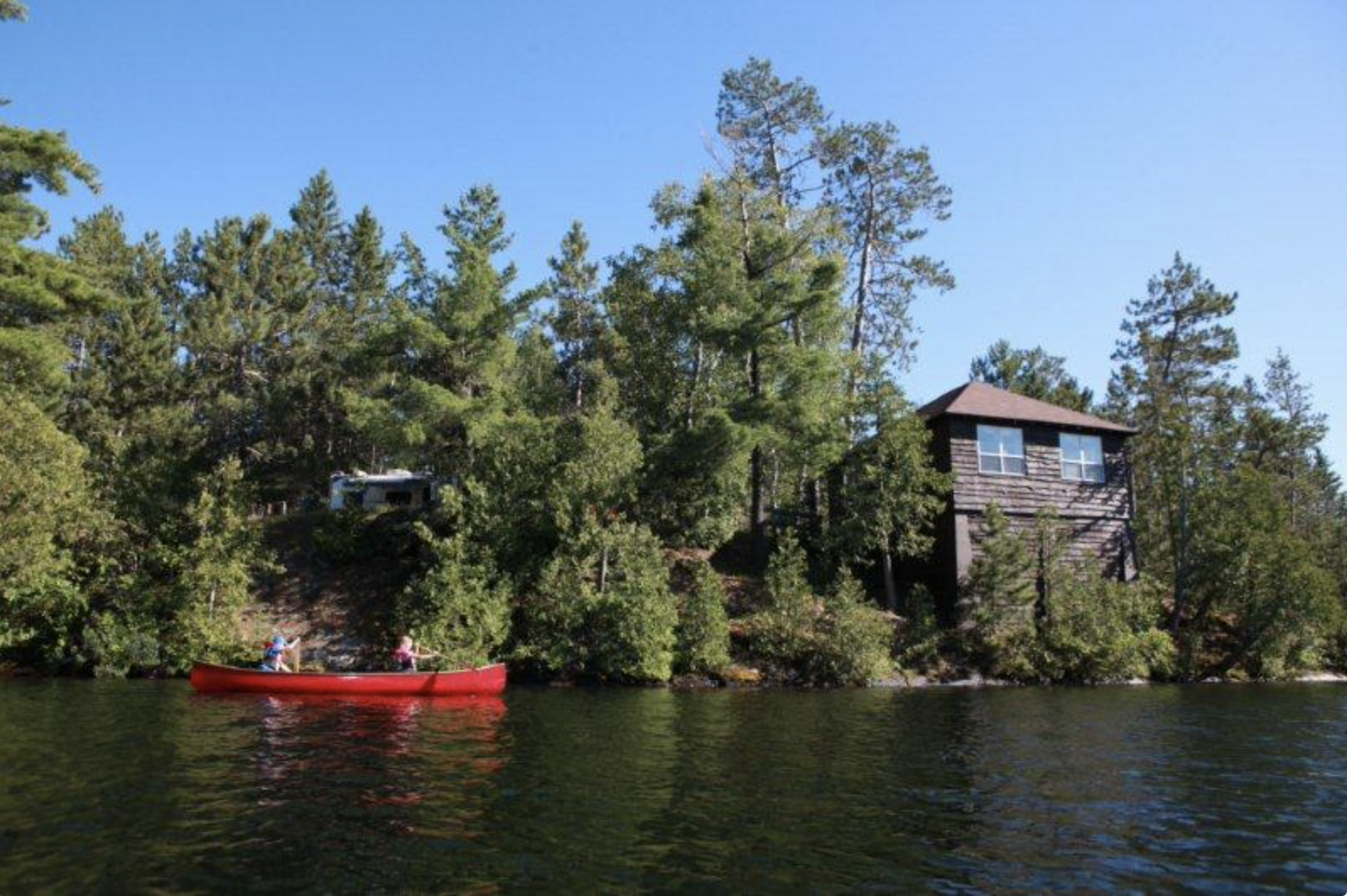 a pretty cabin tucked into the dense green forest on a rocky lake shore, under a brilliant blue sky. A red canoe is floating on the lake near the cabin.