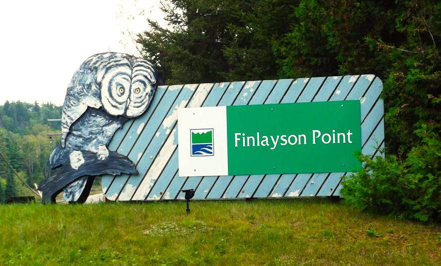 A large green, blue and white wooden Ontario Parks sign that reads "Finlayson Point", on a large, rectangular slatted-wood background with a huge painted owl picture on the left-hand side. There are green grass and trees all around. 