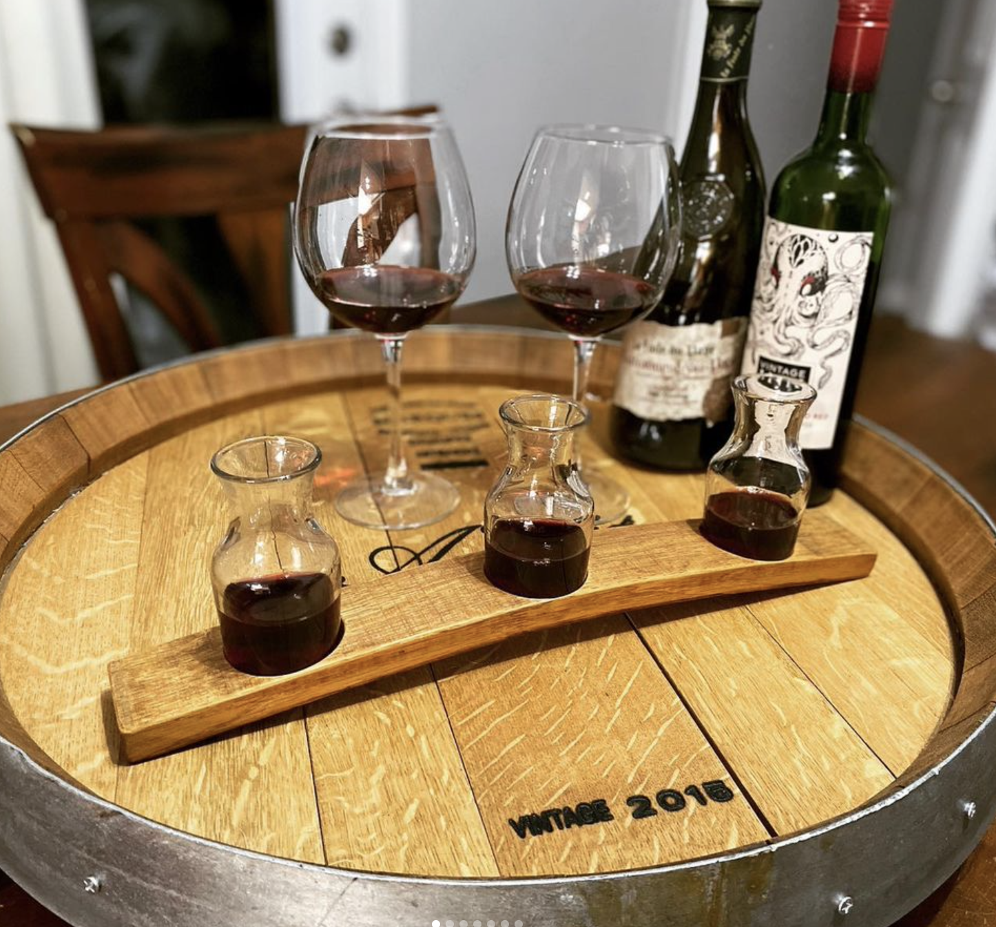 A wooden flight board holding 3 small carafes of wine on top of a wooden wine cask, with wine bottles and glasses in the background.