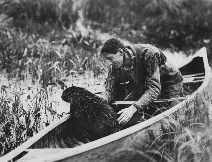 a black and white photo of the conservationist Grey Owl sitting in a canoe with a beaver, floating in a marsh. The man appears to be talking gently to the beaver and has his hand on its back.