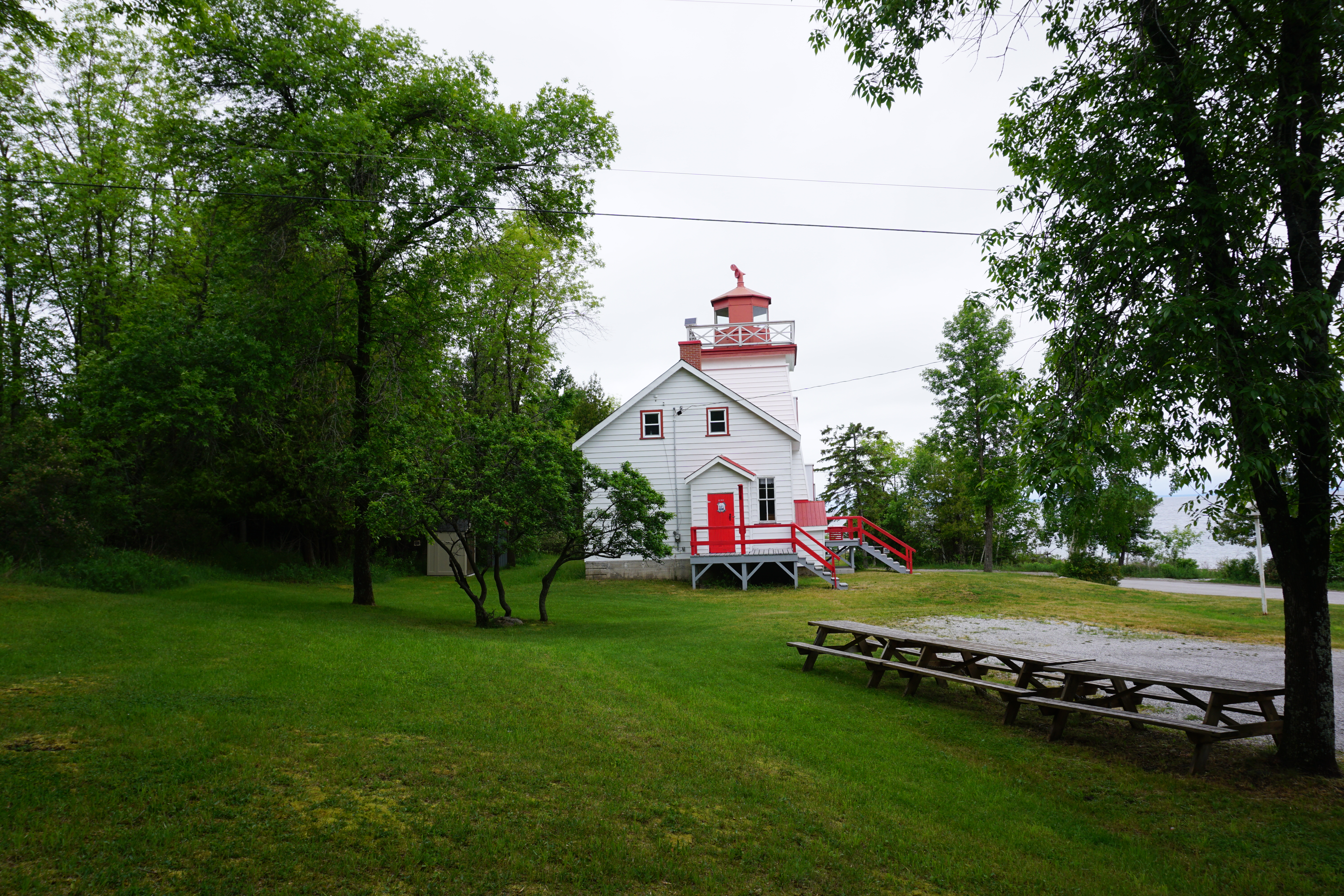 The Jant Head Lighthouse; a short red and white lighthouse on the edge of Gore Bay, surrounded in green grass and trees.