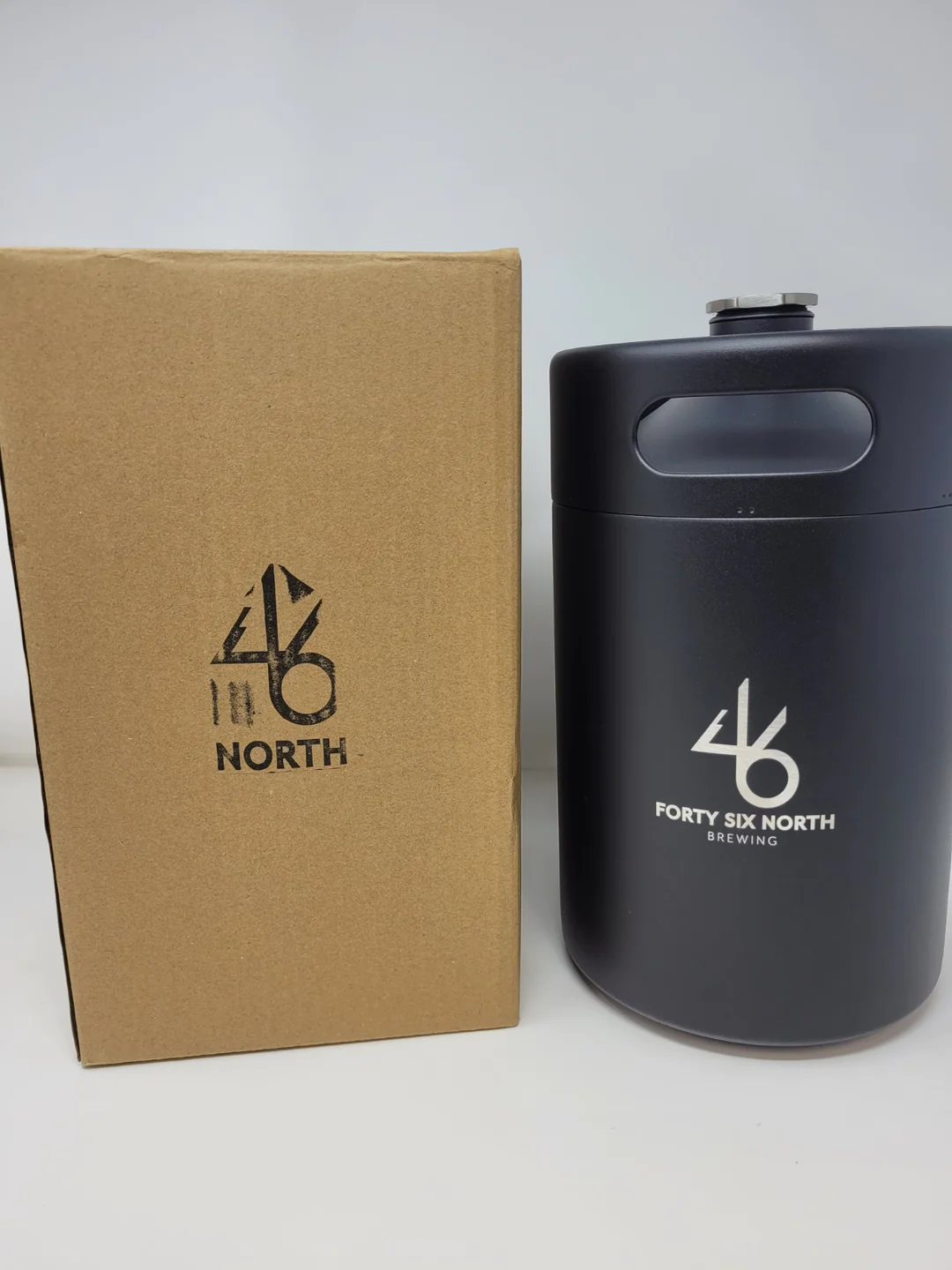 a black painted stainless steel growler next to a brown cardboard box of the same size, both printed with a 46 North logo.