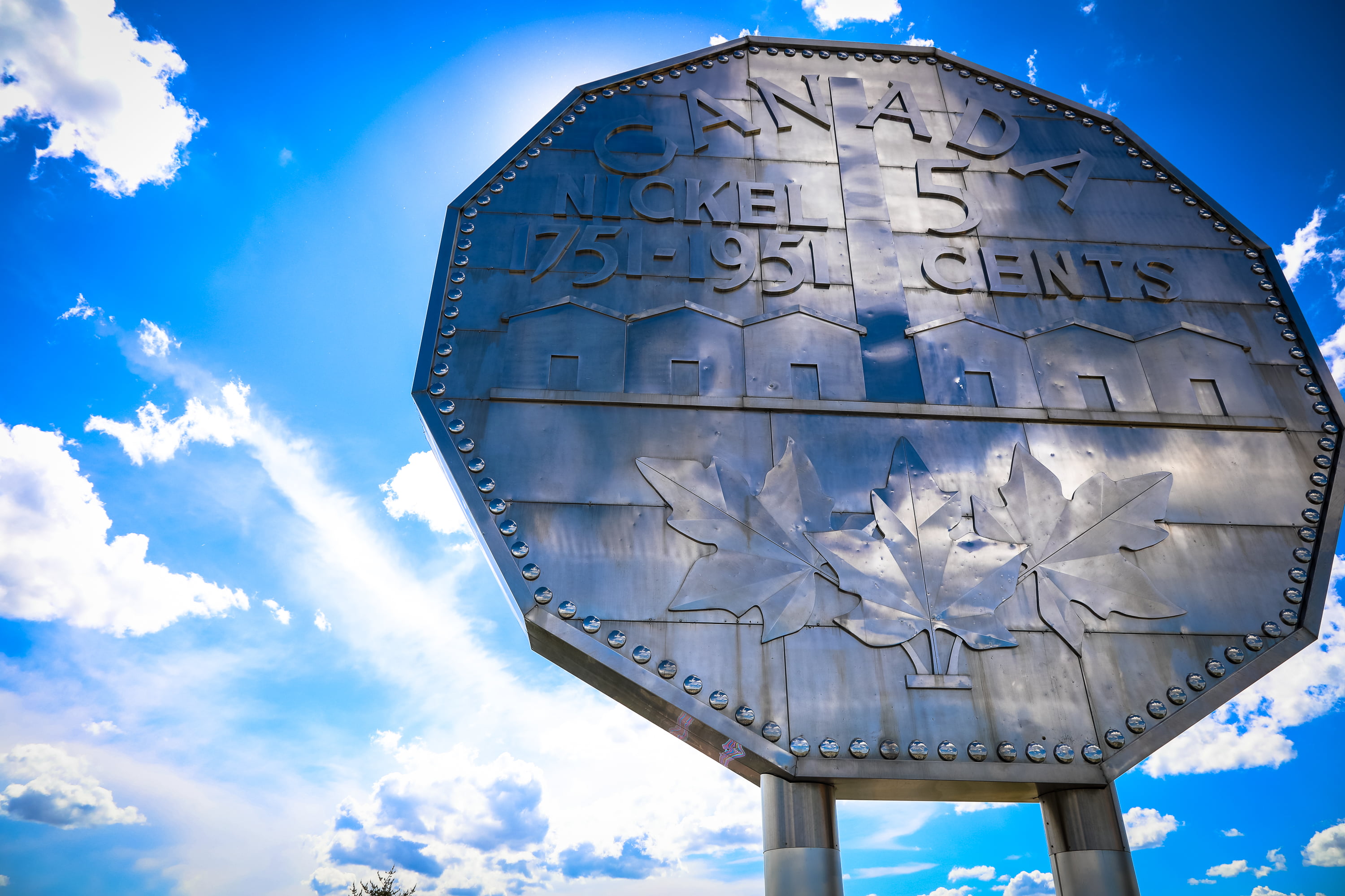 a low shot of Sudbury's Big Nickel monument, standing tall above the camera in front of a brilliant blue sky striped with white clouds.