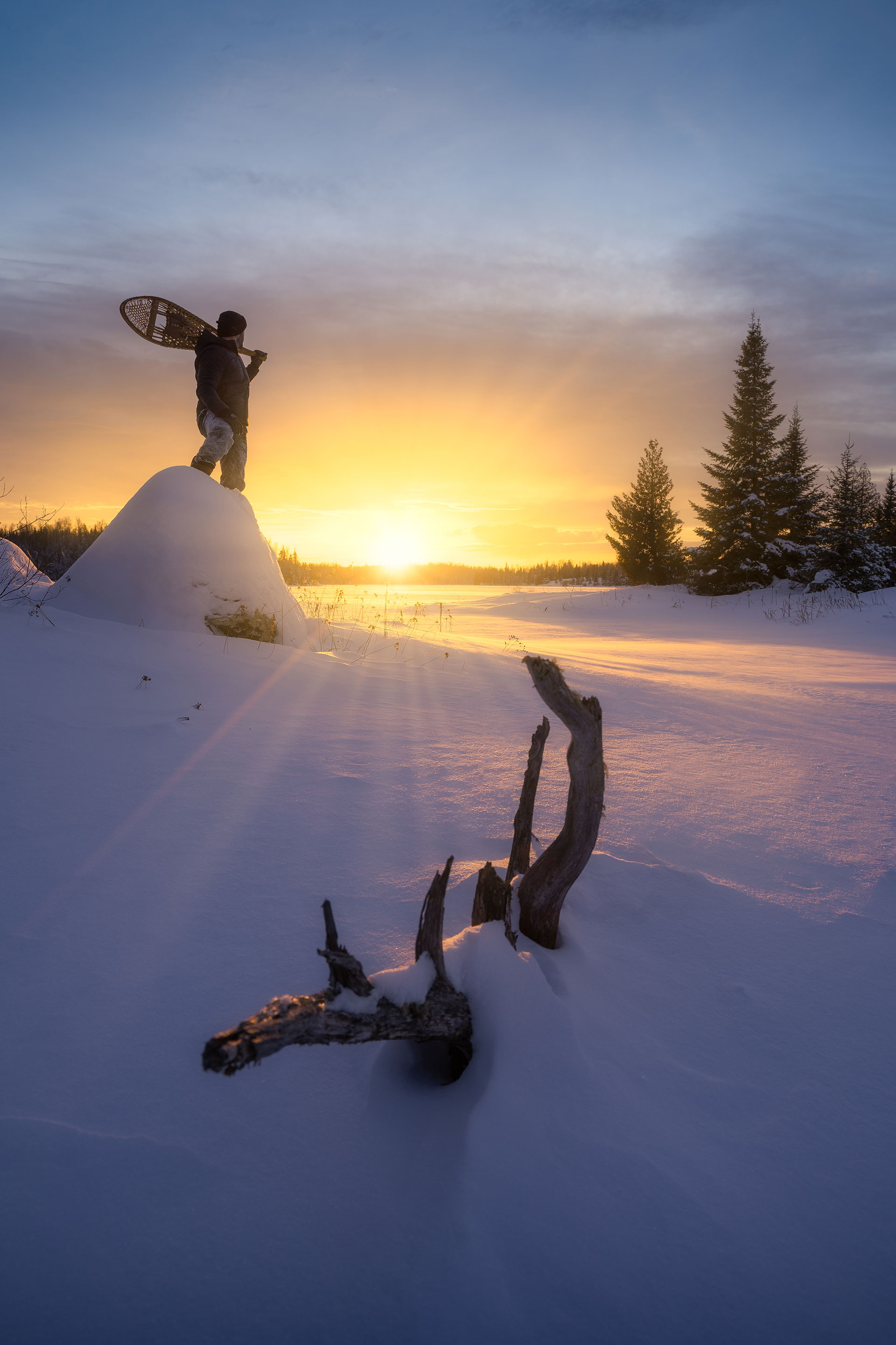 person stands on mound of snow at sunrise while holding snowshoes