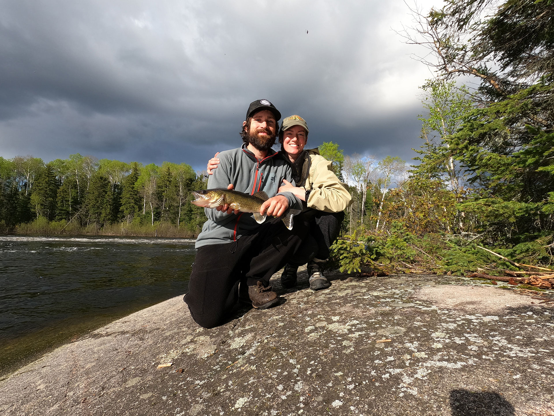 man and woman pose on shoreside rock cap with fish they caught
