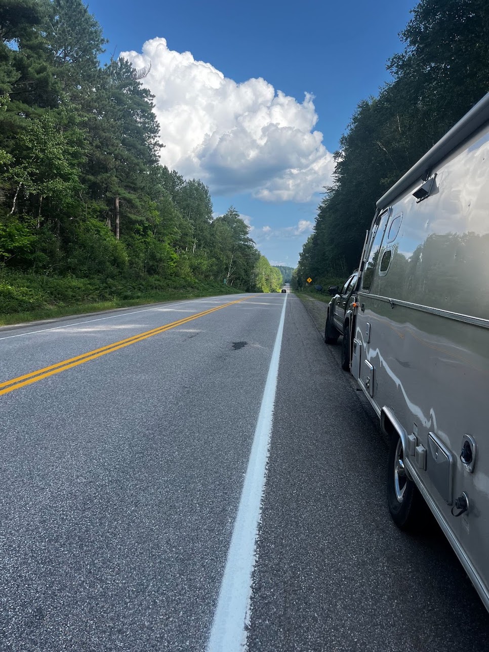 a low shot of the side of a shiny Airstream trailer rolling along an empty highway, which stretches out into the distance. There are walls of green forest on both sides and a bright blue sky overhead.