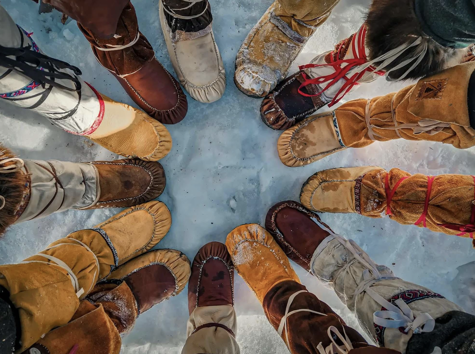 A bunch of feet wearing moccasins placed in a circle in the winter snow. 