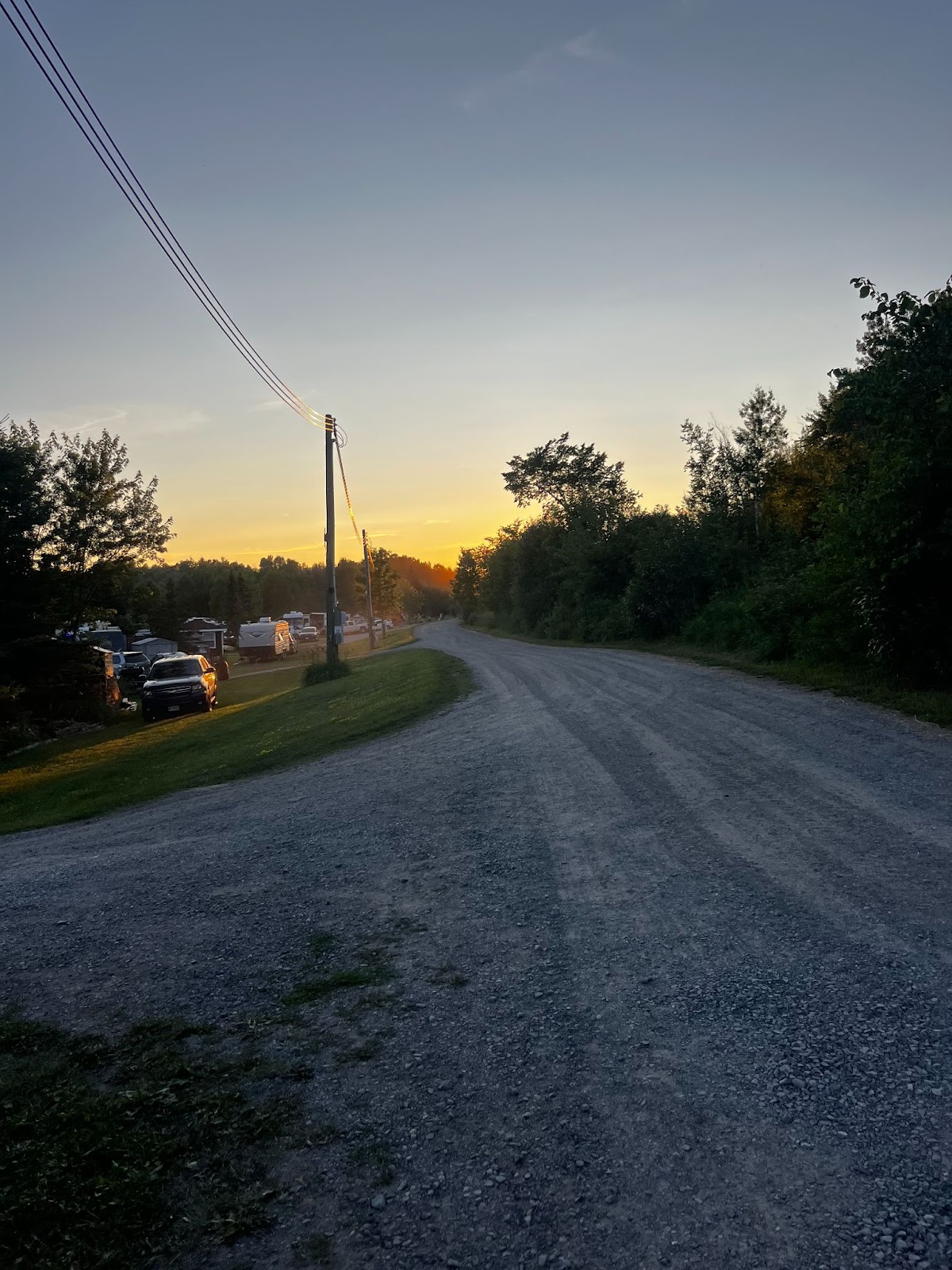 a gravel campground road at dusk, with dark, thick forest on the right and rows of cars and RVs parked in a green field on the left , under a set of power lines. The sky is getting dark and there is the end of an orange sunset in the distance.
