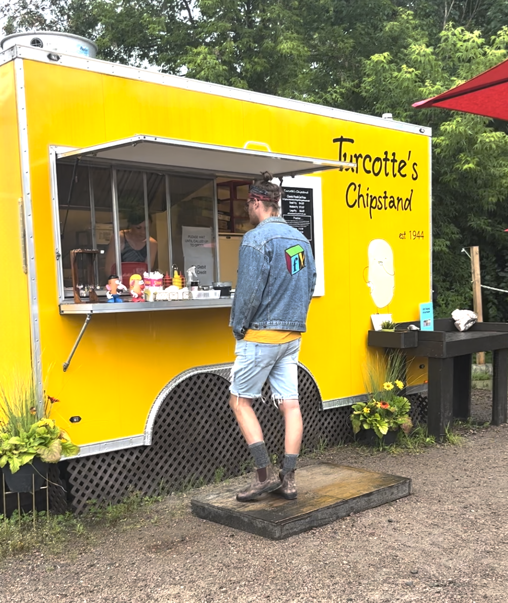 a man visiting a bright yellow mobile Turcotte's Chip Stand, parked in front of a green forest.