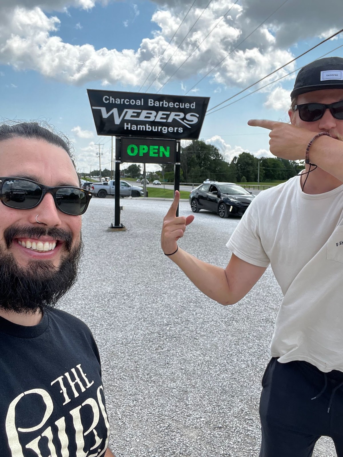 two people smiling and pointing at a roadside sign that reads "Charcoal Barbequed Webers Hamburgers, Open"