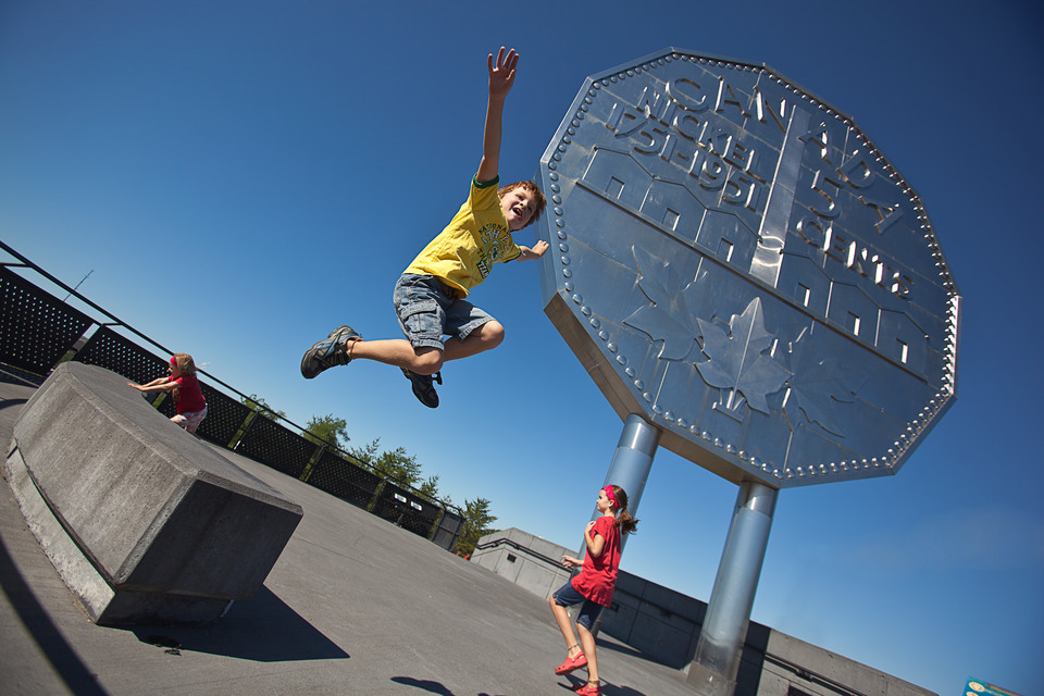 2 kids smiling, jumping and running underneath the Sudbury Big Nickel monument, which towers above them against a bright blue sky.