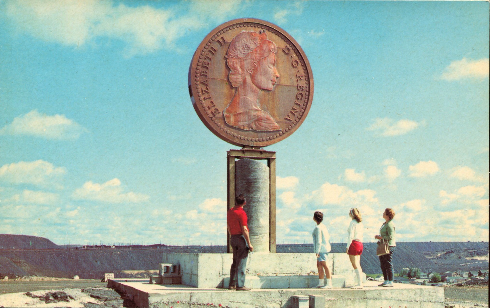 an aged photograph of 4 tourists looking up at the Penny monument, formerly in Sudbury; a giant replica penny set on a pillar, in front of a blue sky.