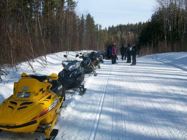 a group of people talking on a forested snowmobile trail, their snowmobiles parked in a line.
