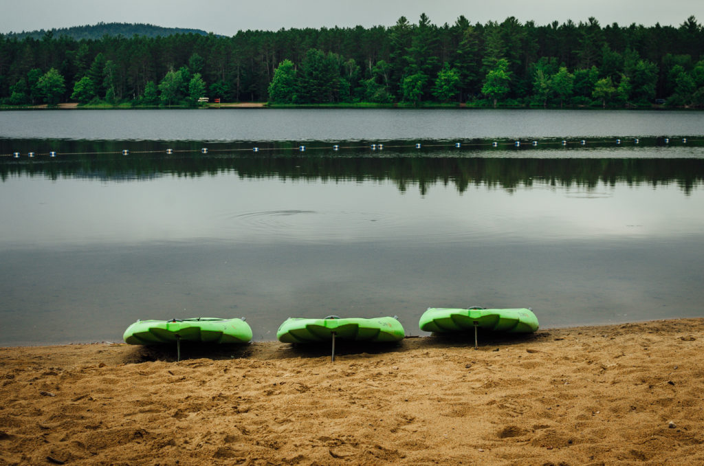 the beach at Samuel de Champlain Provincial Park campground; a large sandy beach with a still, glassy lake and dense forest on the opposite bank. There are 3 green inflatable water boards lined up on the beach.
