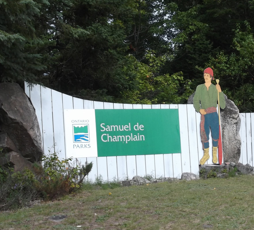 A white and green wooden Ontario Parks sign that reads "Samuel de Champlain", with a wooden Voyageur cutout standing at one side. There are green grass and trees all around.