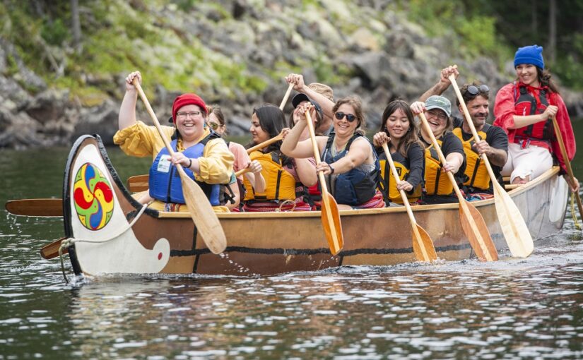 a row of smiling people paddling a large birchbark canoe on a river. The end two appear to be paddling instructors, dressed as voyageurs.