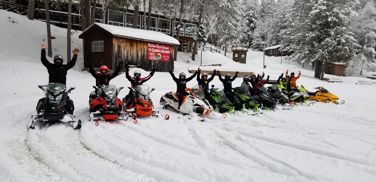 A line of people on snowmobiles, all with their arms raised in the air, parked in a snowy driveway in front of wooden building with a sign that reads "Mattawa River Resort"