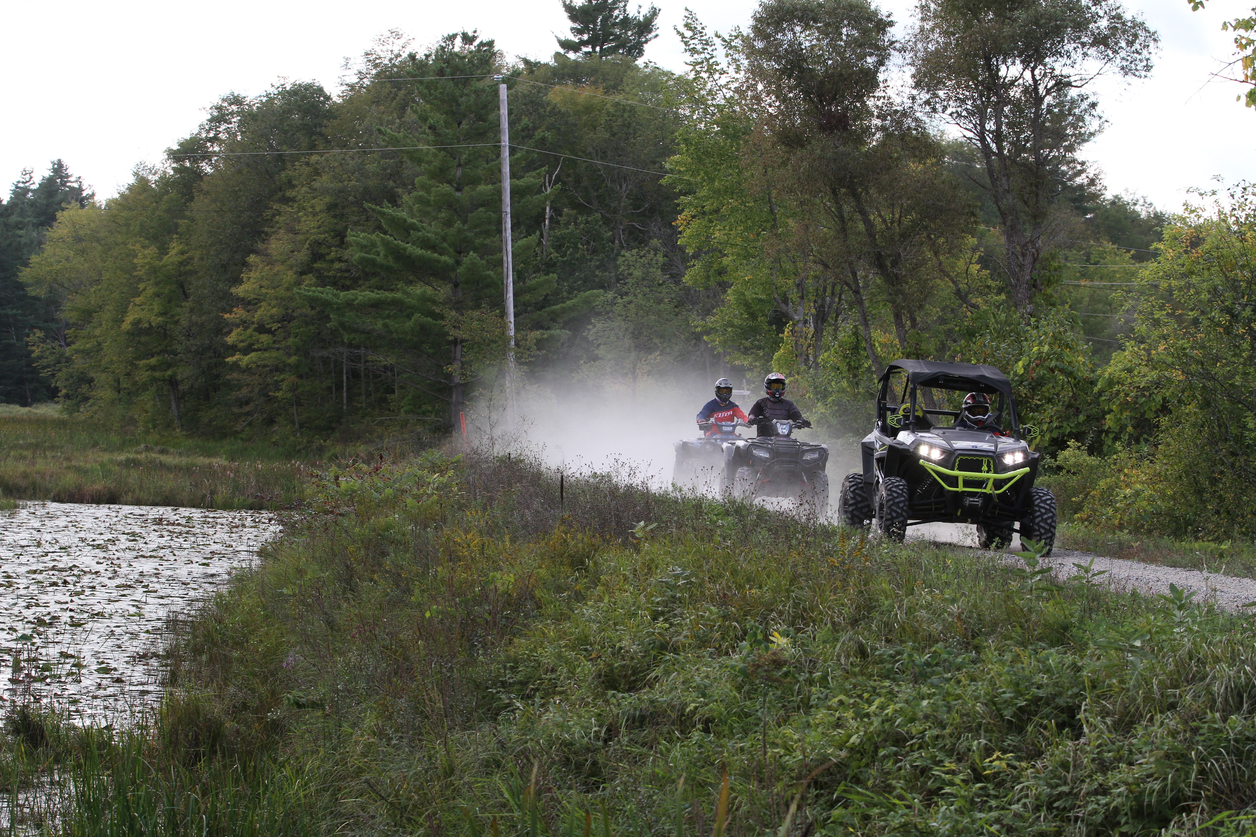 3 ATVs driving down a dusty trail surrounded by green forest, grass and a lake to the left.