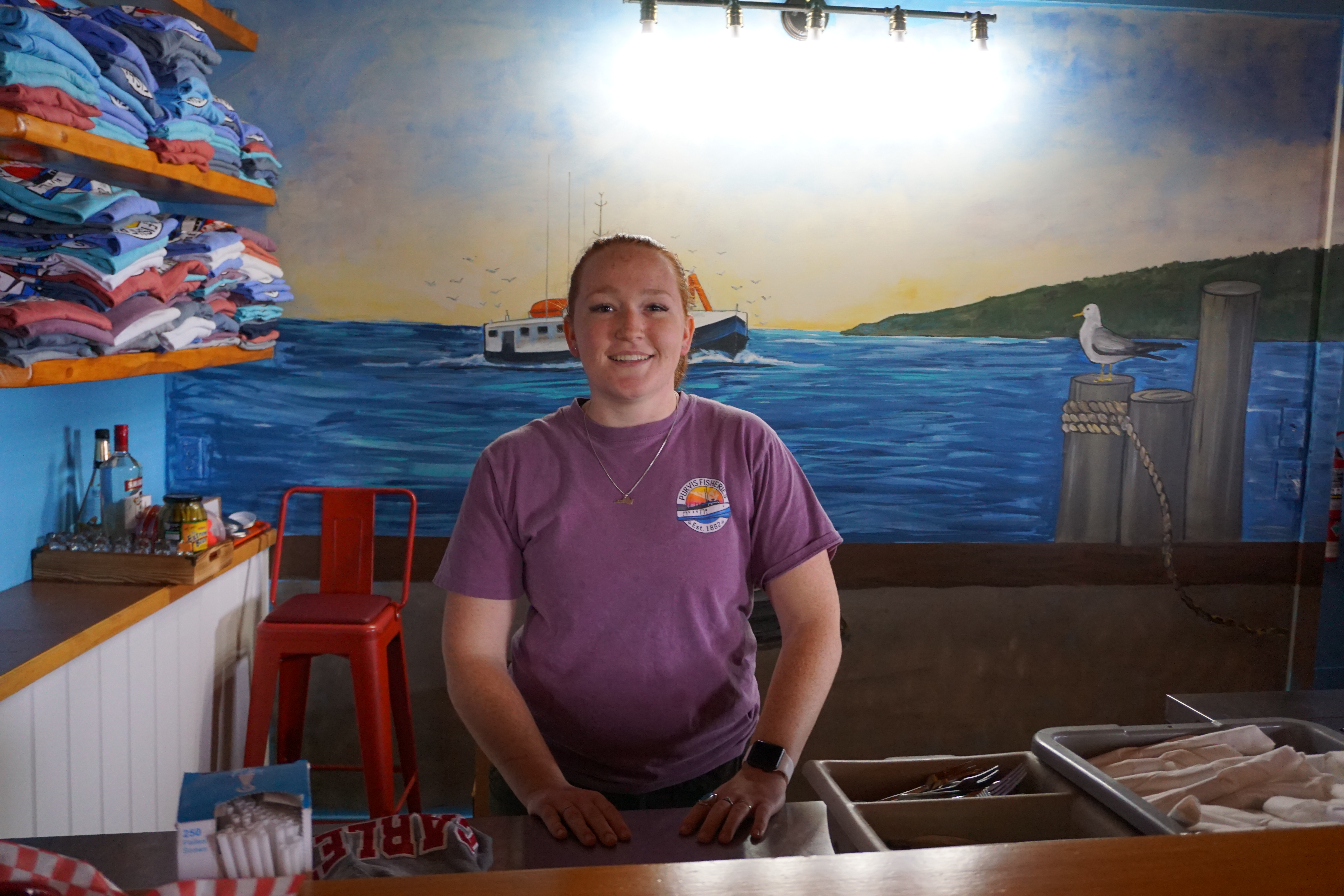 Owner Avery Sheppard smiling behind the counter of Purvis Fish & Chips, a mural of a blue harbour and fishing boat on the wall behind her.