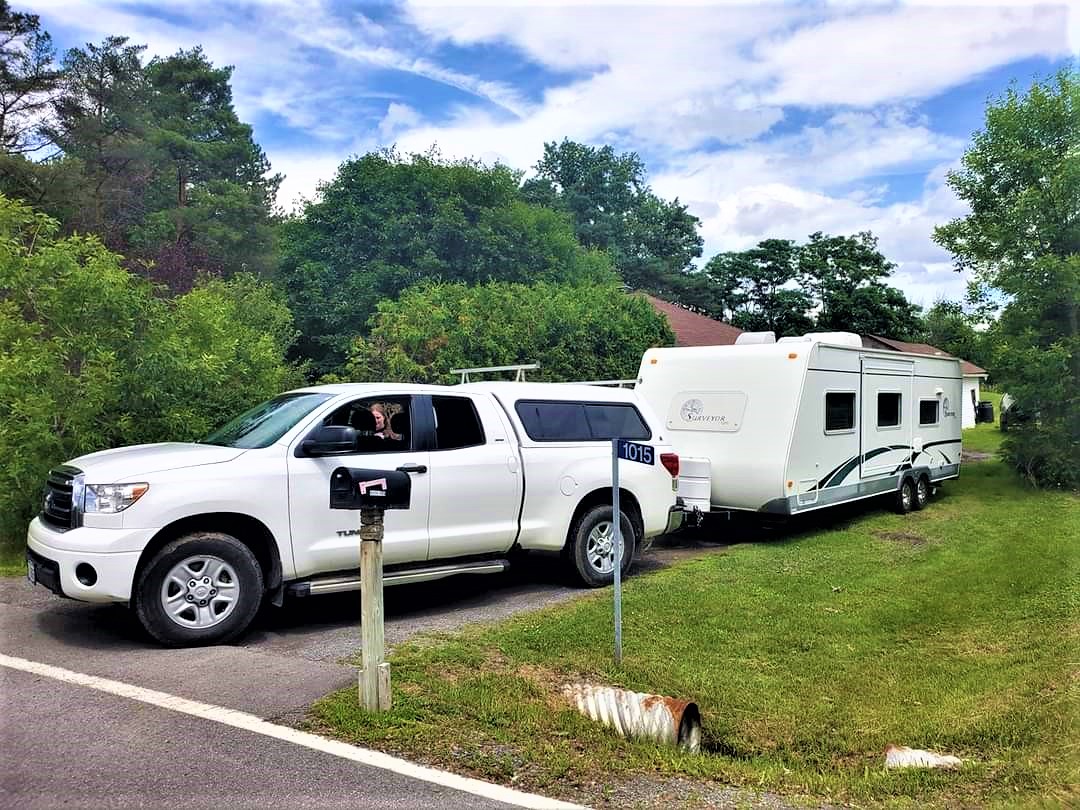 A white truck towing a large white RV camper, parked in a residential driveway. It is surrounded in lush summer greenery under a blue sky.