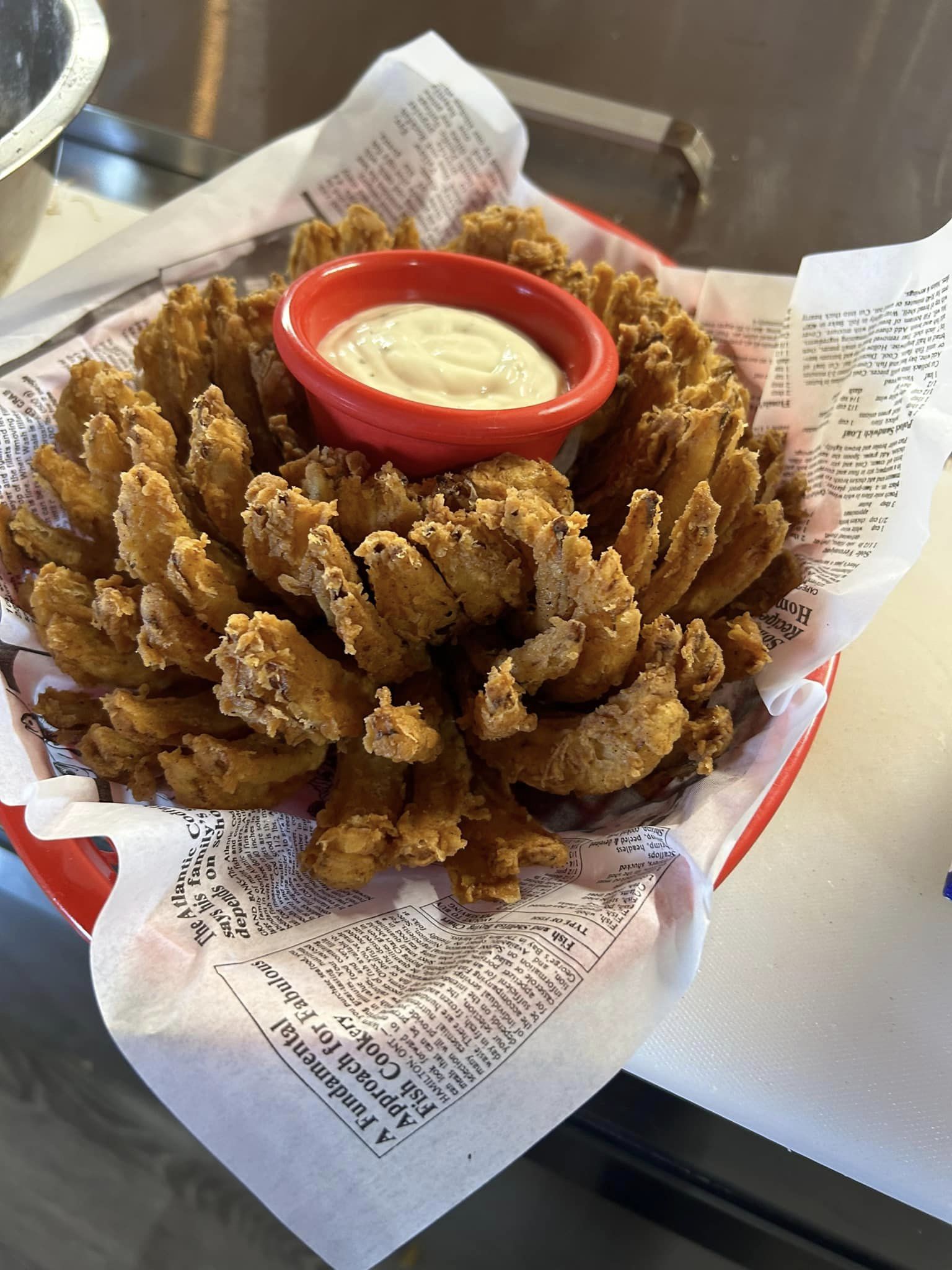 a deep fried bloomin' onion served in a paper lined basket with a bowl of dip in the center.
