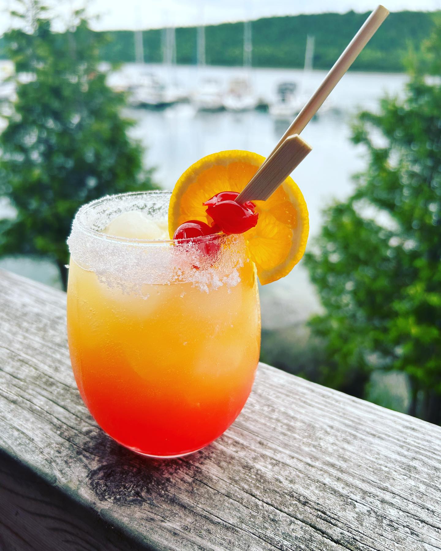 an orange and red cocktail decorated with a sugared rim, cherries and a lemon wedge, placed on a wooden rail. Summer trees and a boat-filled harbour can be seen in the background. 