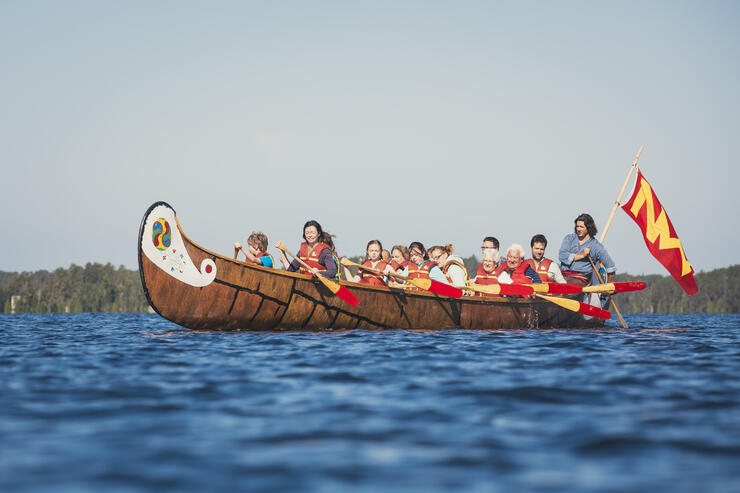 People paddling a big wooden canoe on a blue lake.