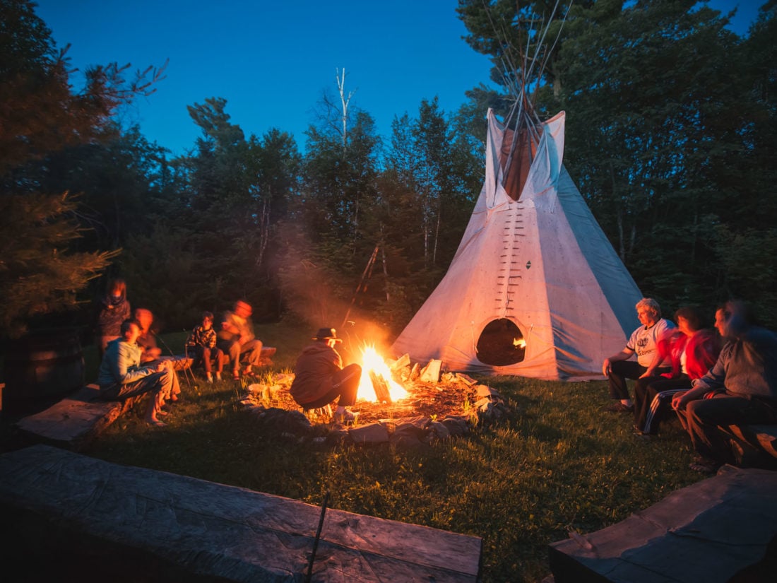 A number of people sit around a campfire in front of a white teepe at dusk. 