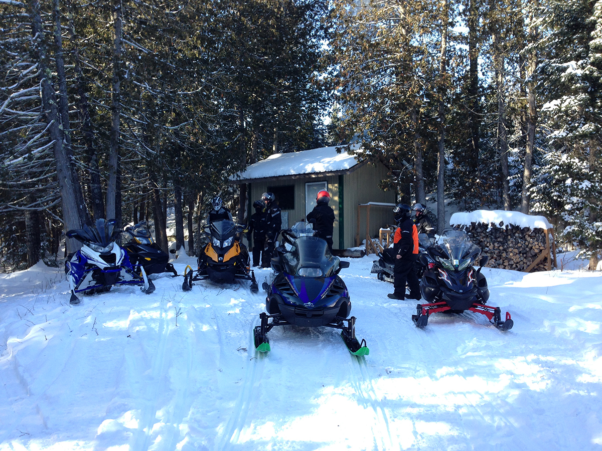 a group of snowmobilers parked at a warm-up shack along a forested snowmobile trail