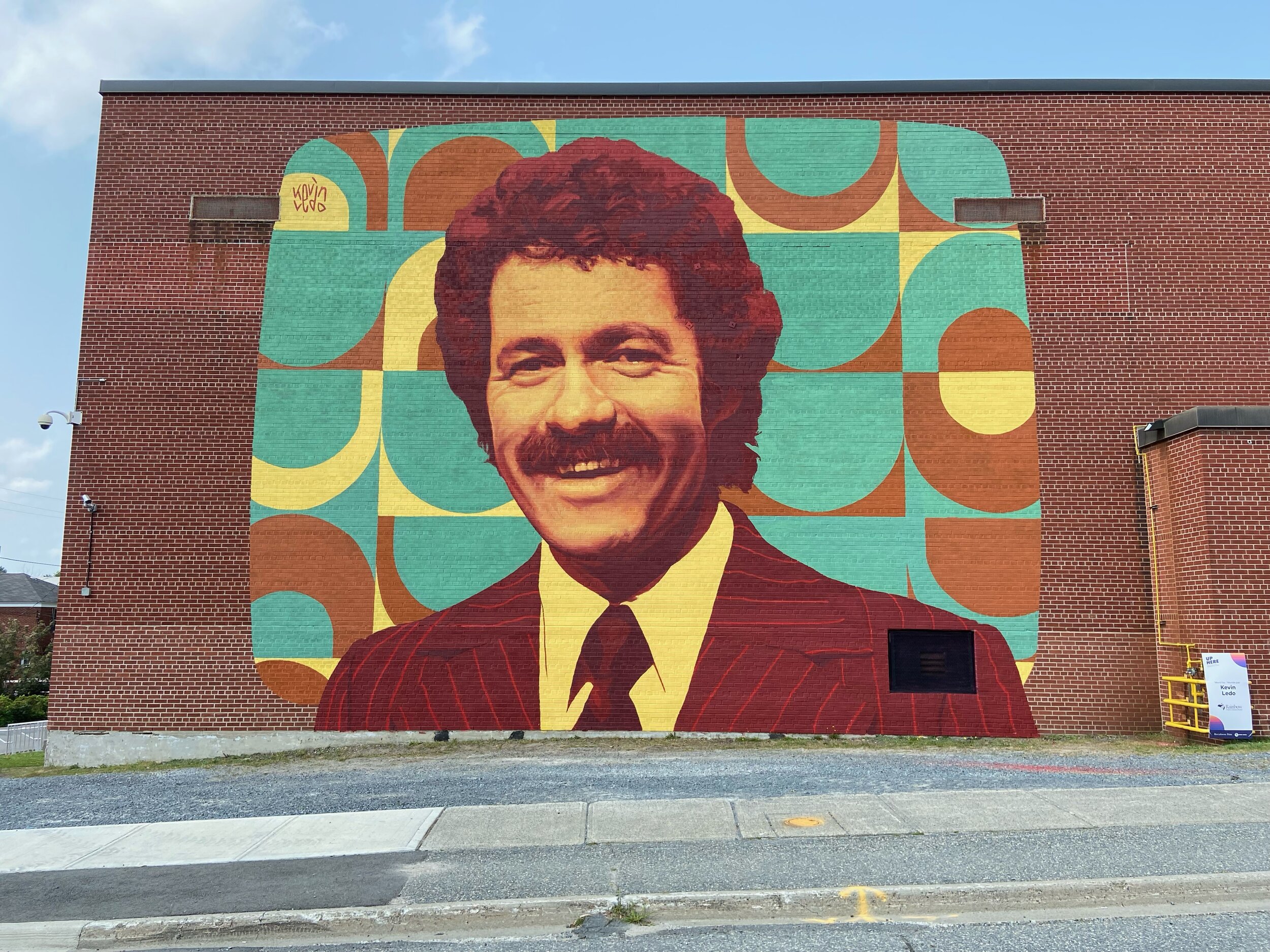 A large outdoor mural of Alex Trebek; a smiling man with brown hair and a moustache wearing a brown suit and tie in front of a turquoise and yellow patterned background.
