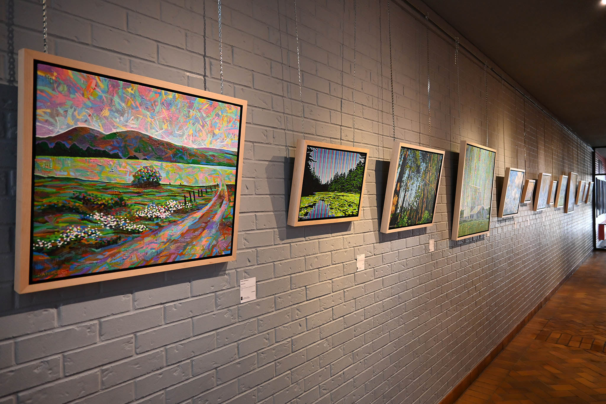 Interior of the Art Gallery of Sudbury; a white brick wall in a dimmed gallery, hung with a long row of beautiful nature paintings under spotlights.
