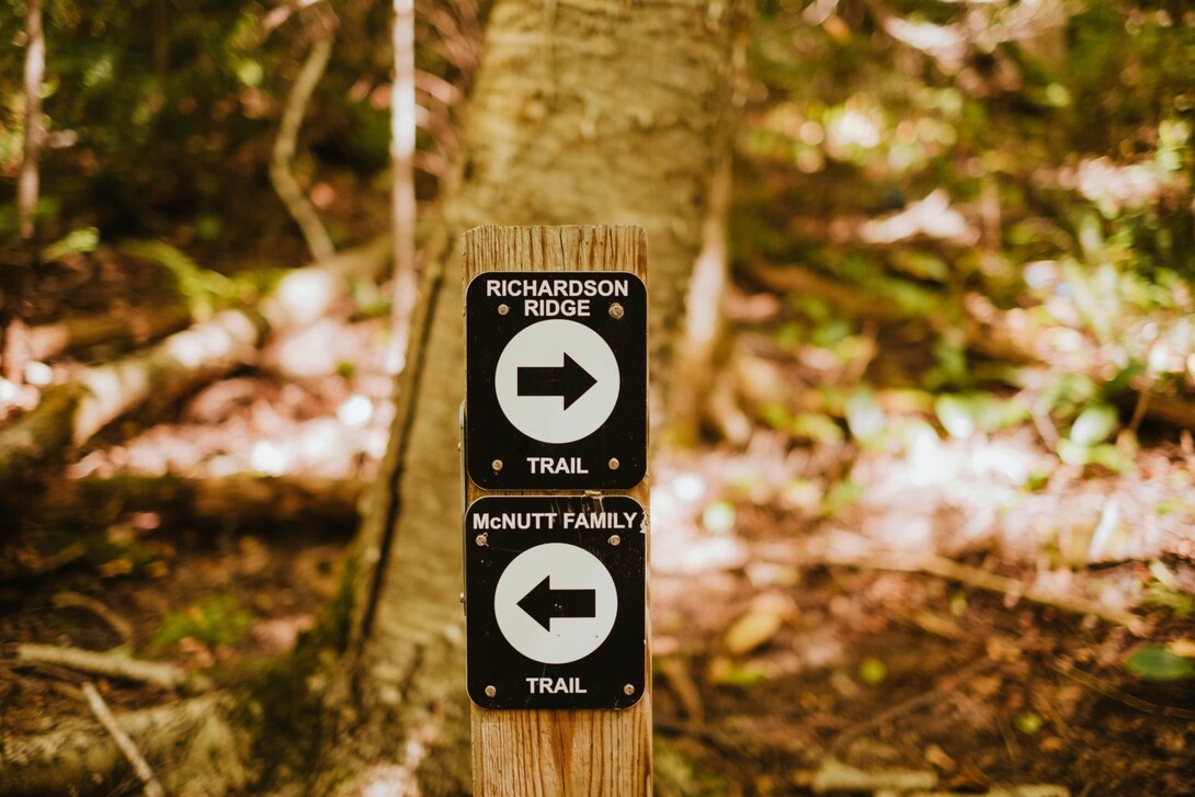 Two black and white directional trail marker signs attached to a tree on a forest path; one reads "Richardson Ridge Trail", the other, "McNutt Family Trail"