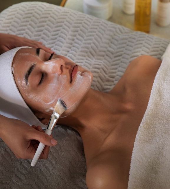 a woman with her hair wrapped in a white towel and a relaxed look on her face receives a facial treatment at a spa