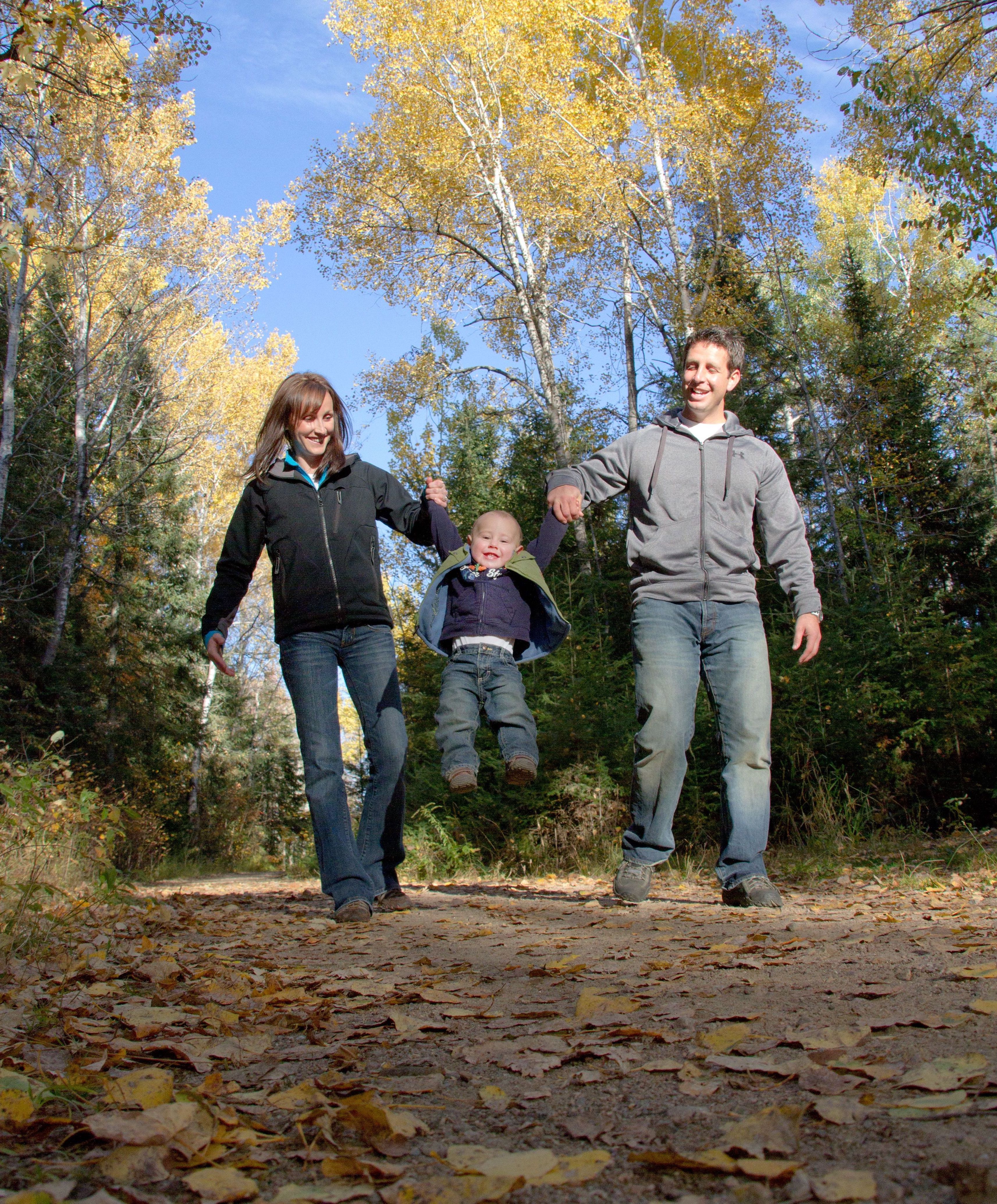 a mother and father each hold the hand of a toddler, smiling as they swing the laughing child off the ground by the hands as they walk on a forest trail. The forest leaves are yellow, green and orange and the sky is bright blue.