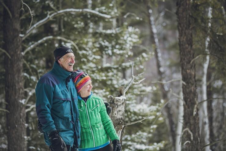  an older couple dressed in winter gear, smiling widely as they look at the snow-covered forest around them