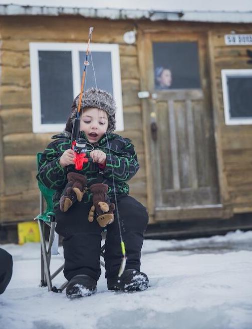 a small child sitting in a miniature folding chair, gasping in surprise as they ice fish. A wooden fishing shack is in the background.