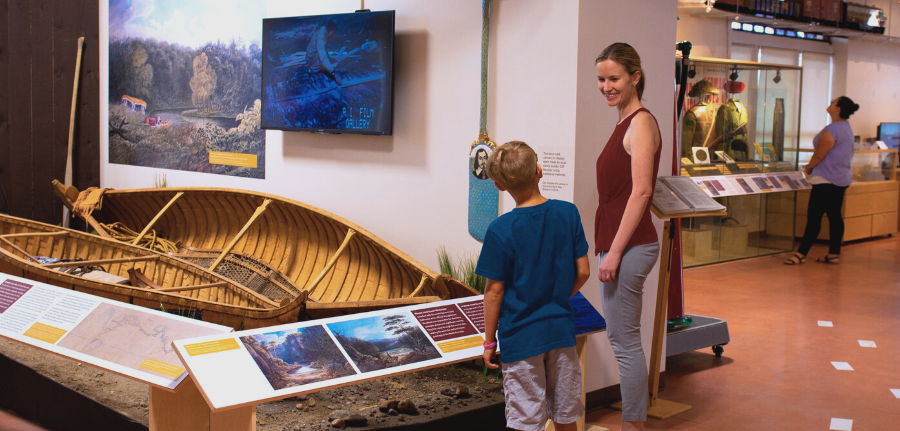 a woman and boy talking to each other and smiling as they look at a canoe exhibit at the North Bay Museum