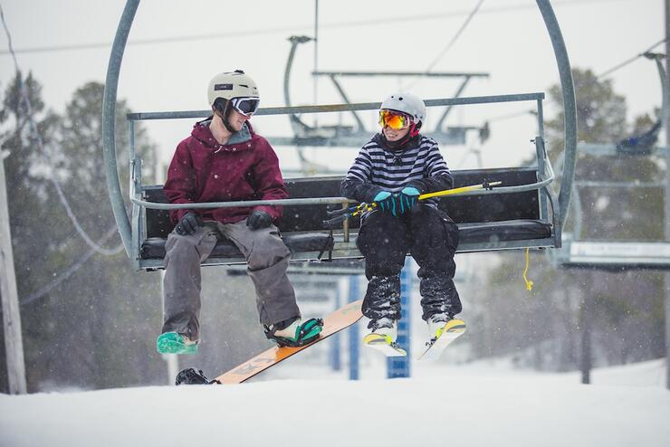 Two young people talking and smiling while riding a ski lift with their equipment on a snowy day