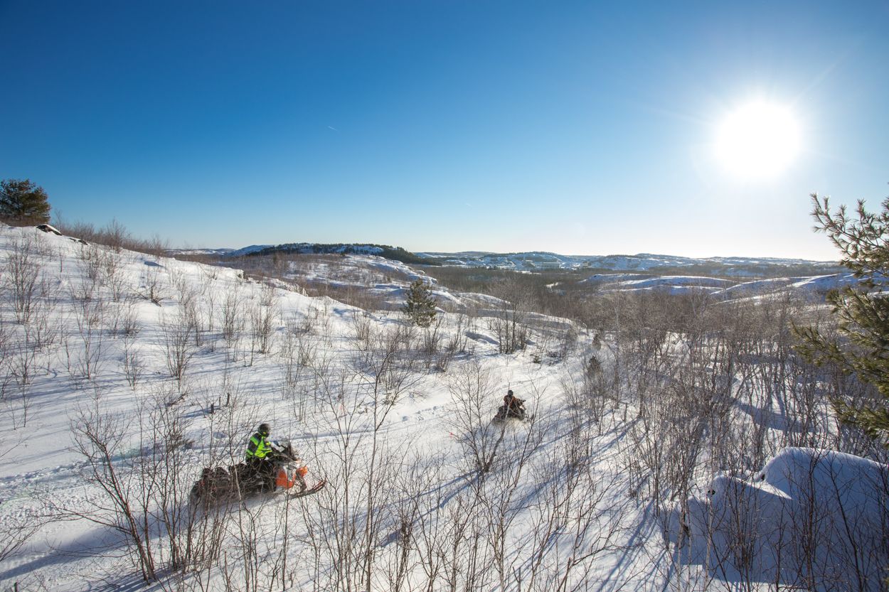 a high shot of two snowmobilers riding through a snowy valley below, the hills covered with saplings and forest in the distance, all under a blazing sun and bright blue sky.