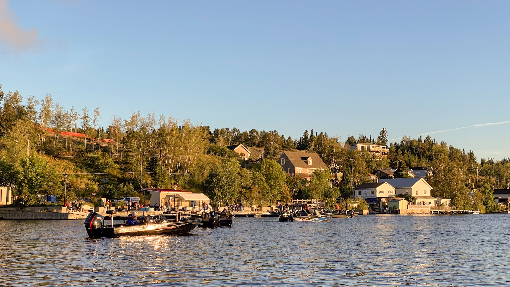 Red Lake; motor boats float in calm blue water next to docks on a green, forested lakeshore dotted with houses. Golden late afternoon sun shines in a hazy blue sky. 