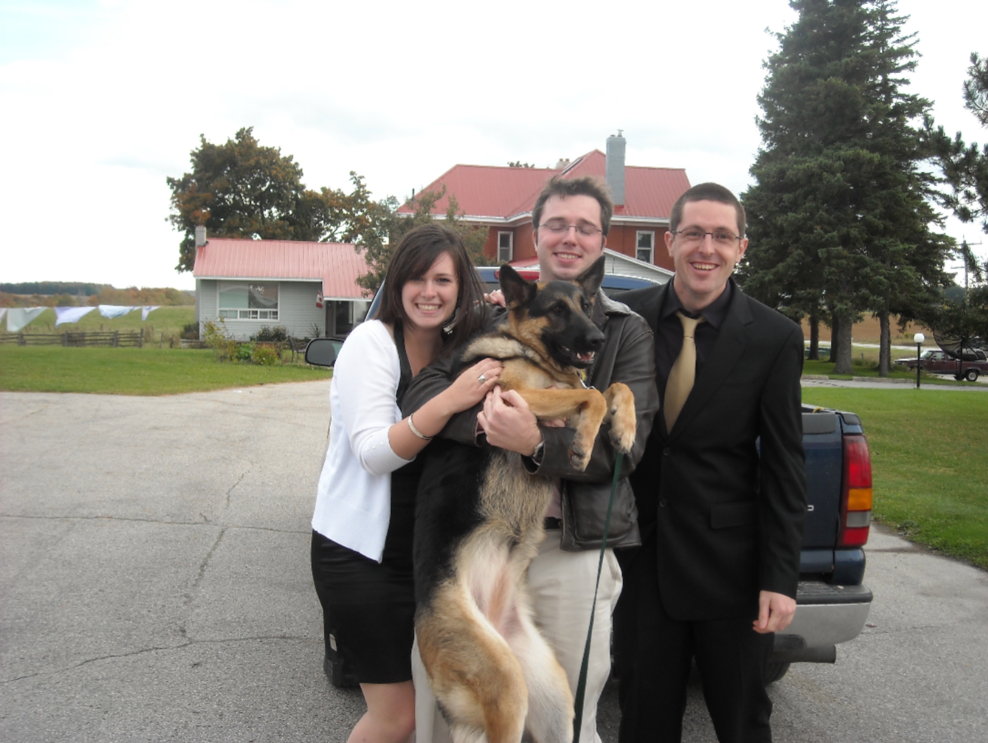 A young woman and two young men standing outside in formal dress, smiling. The man in the centre is hugging a large german shepherd to his chest, raising the dog to standing position. A large, red-roofed lake house, lake and green trees are in the background.