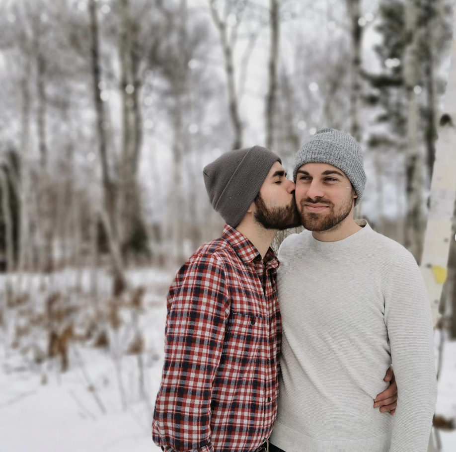 Two bearded men in long sleeve tops and hats with snow and trees. 