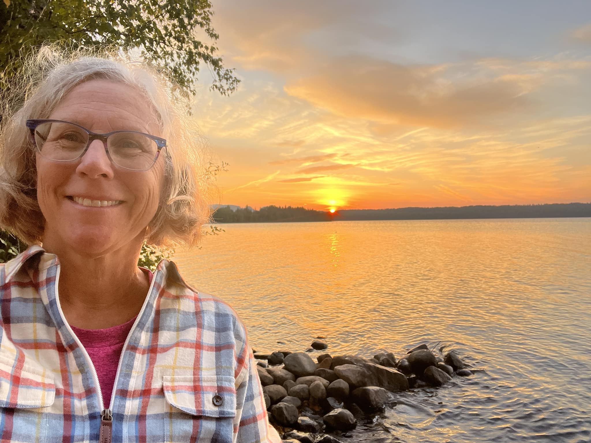 Laura MacGregor, smiling on a lakeshore in front of a beautiful orange sunset over the water.