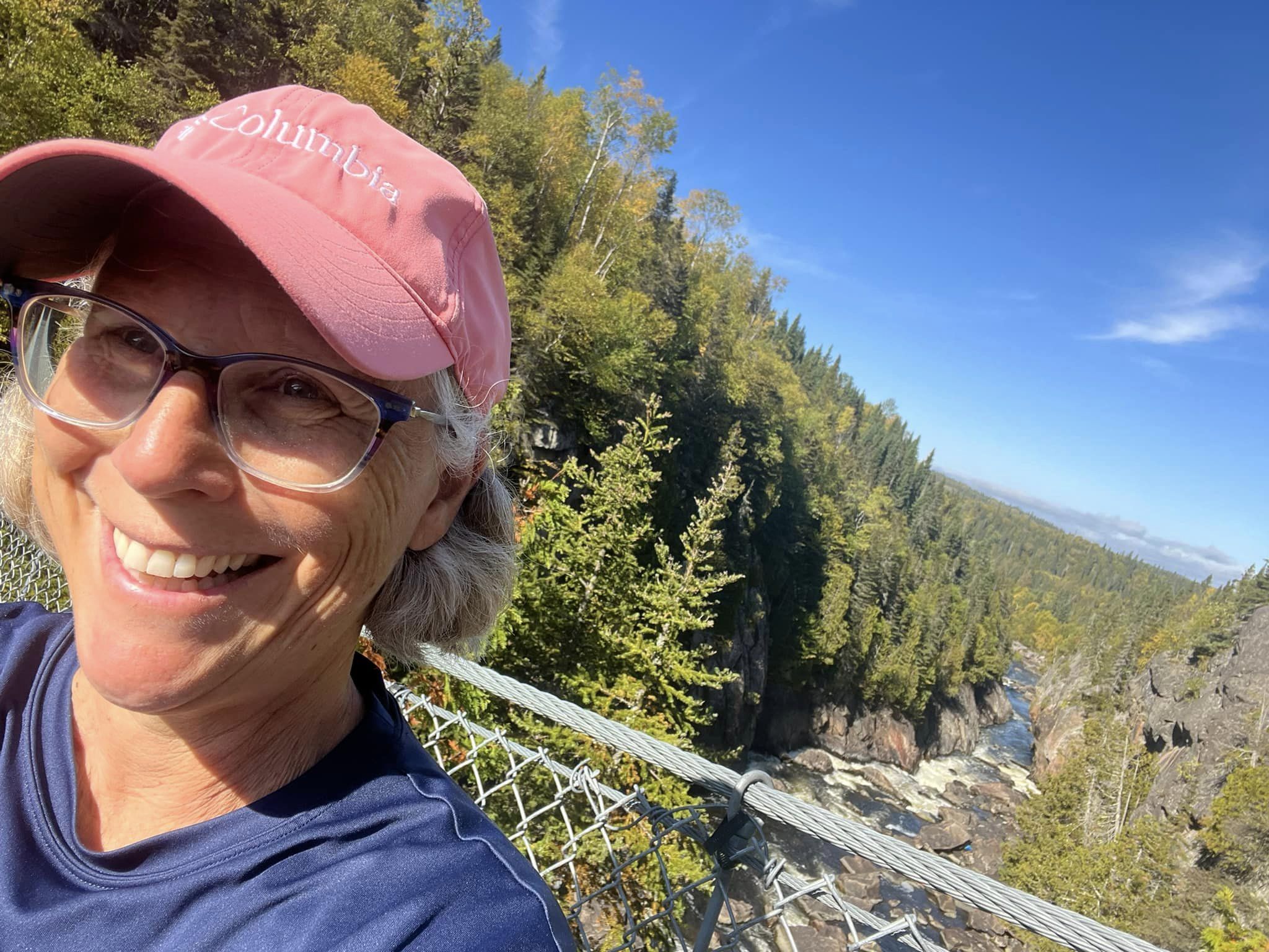 Laura McGregor smiling on a suspension bridge over a rocky riverbed filled with green spruce forest, under a bright blue sky.